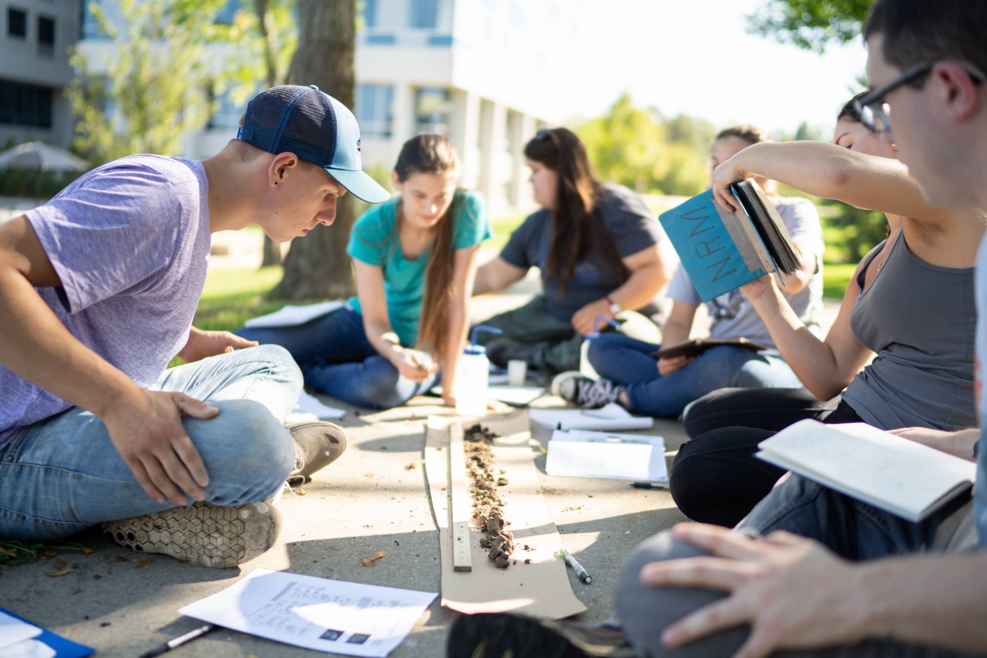 Students sitting on the ground outside gathered around a soil sample and analyzing it